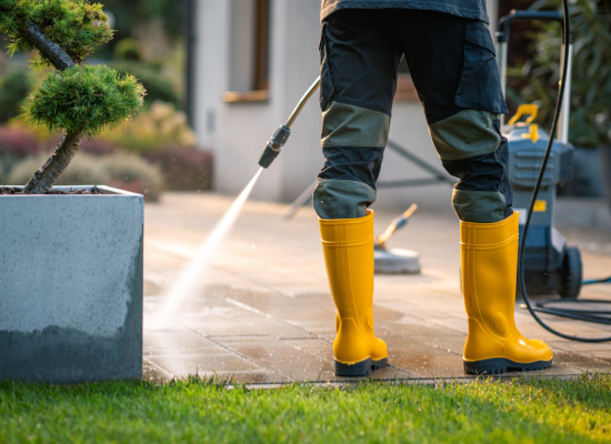 A person wearing yellow rubber boots operates a pressure washer, cleaning a patio area surrounded by greenery. The scene conveys a bright, sunny day in a residential setting.